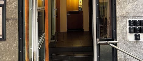 Un área de recepción con puerta de vidrio, un letrero sobre la puerta, una escalera que da a su interior y una pequeña maceta exterior con flores.