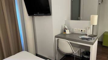 A hotel room with a bed, a desk with a chair, a television, a mirror, a lamp, and a trash bin.