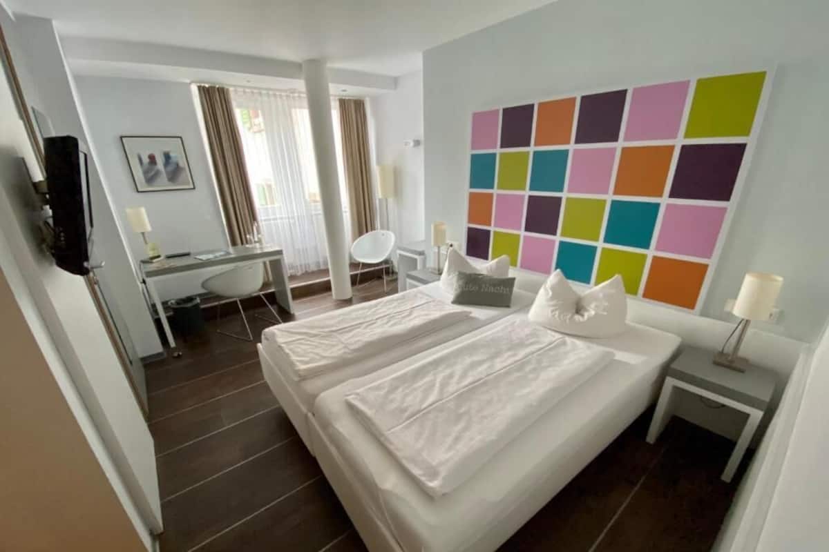 A hotel room with a large bed, a desk, a chair, a colorful wall art, and a small bedside table.