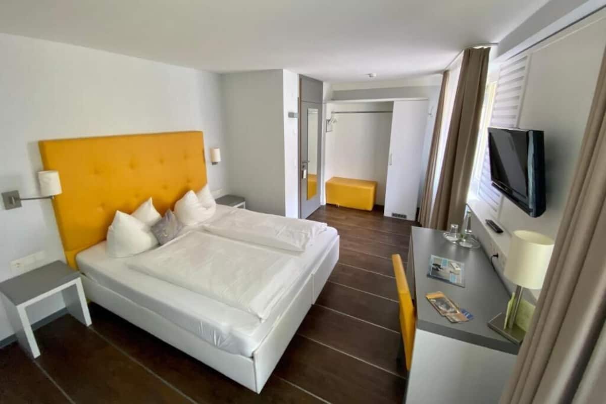 A hotel room with a double bed, a bedside table, a TV, and a small desk with a lamp.