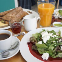 Una mesa con alimentos para el desayuno, incluyendo tostadas, un vaso de zumo de naranja, una taza de té, un bol de sopa y una ensalada con salsa roja.