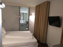 Habitación de hotel con cama, televisor, baño visible a través de un panel de vidrio y una cortina.
