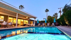 Une piscine avec des chaises longues et des parasols, un patio couvert et des palmiers en arrière-plan.