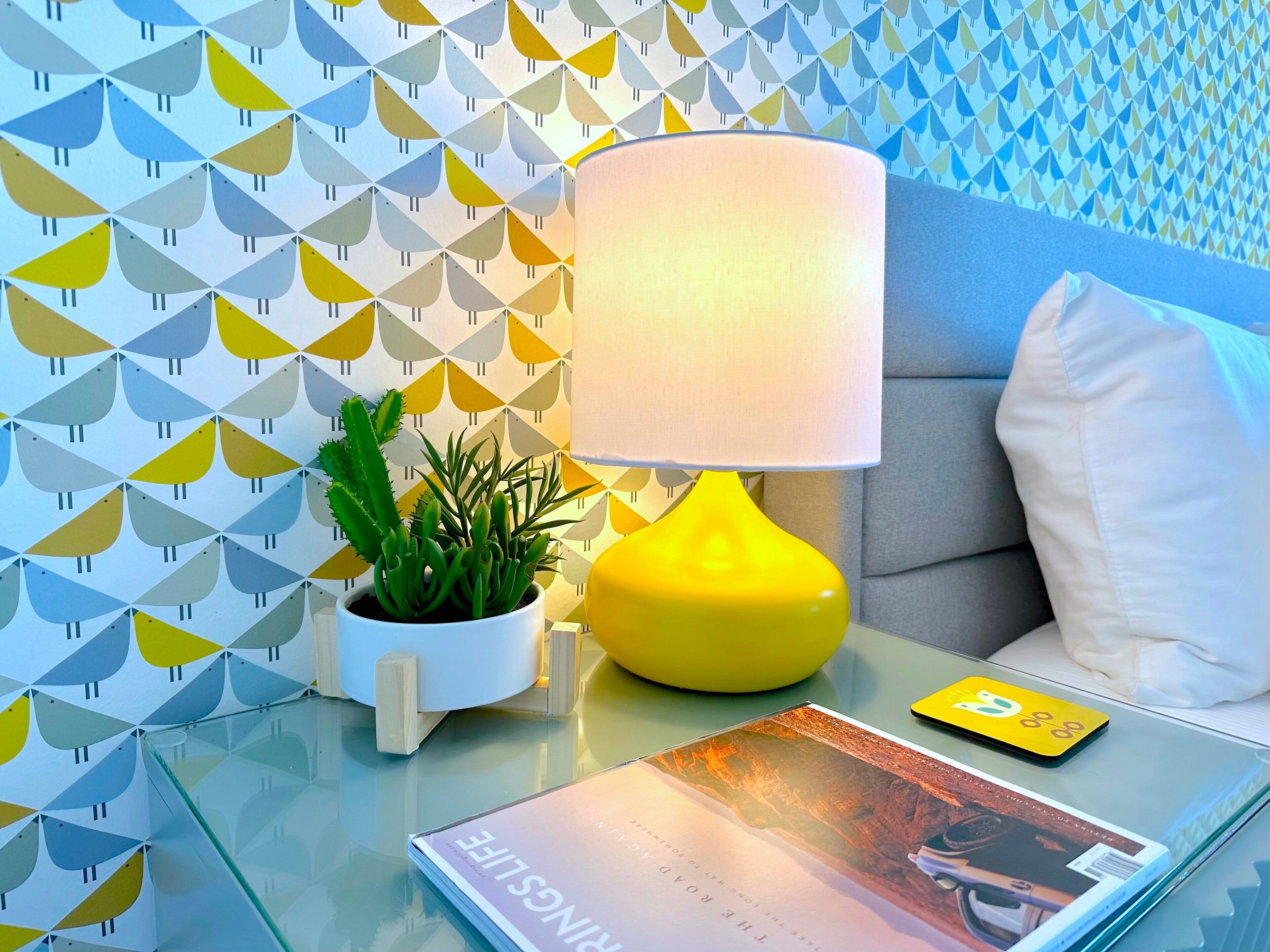 A modern room with a geometric patterned wall, a yellow lamp, a potted plant, a magazine, and a smartphone on a glass table.