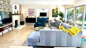 Un salon moderne avec un canapé gris, une table basse et un téléviseur posé sur un meuble. On y trouve également une cheminée, un coin repas et une grande fenêtre donnant sur un paysage verdoyant.