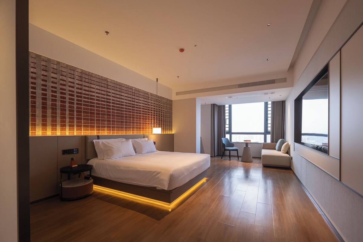 Une chambre d’hôtel moderne dotée d’un grand lit, d’un coin salon avec un fauteuil et une petite table, ainsi que d’une grande fenêtre donnant sur la ville.
