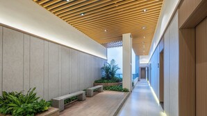 Un pasillo con techo de madera, paredes grises y suelo de baldosas. Se observan bancos y macetas a lo largo de los laterales.