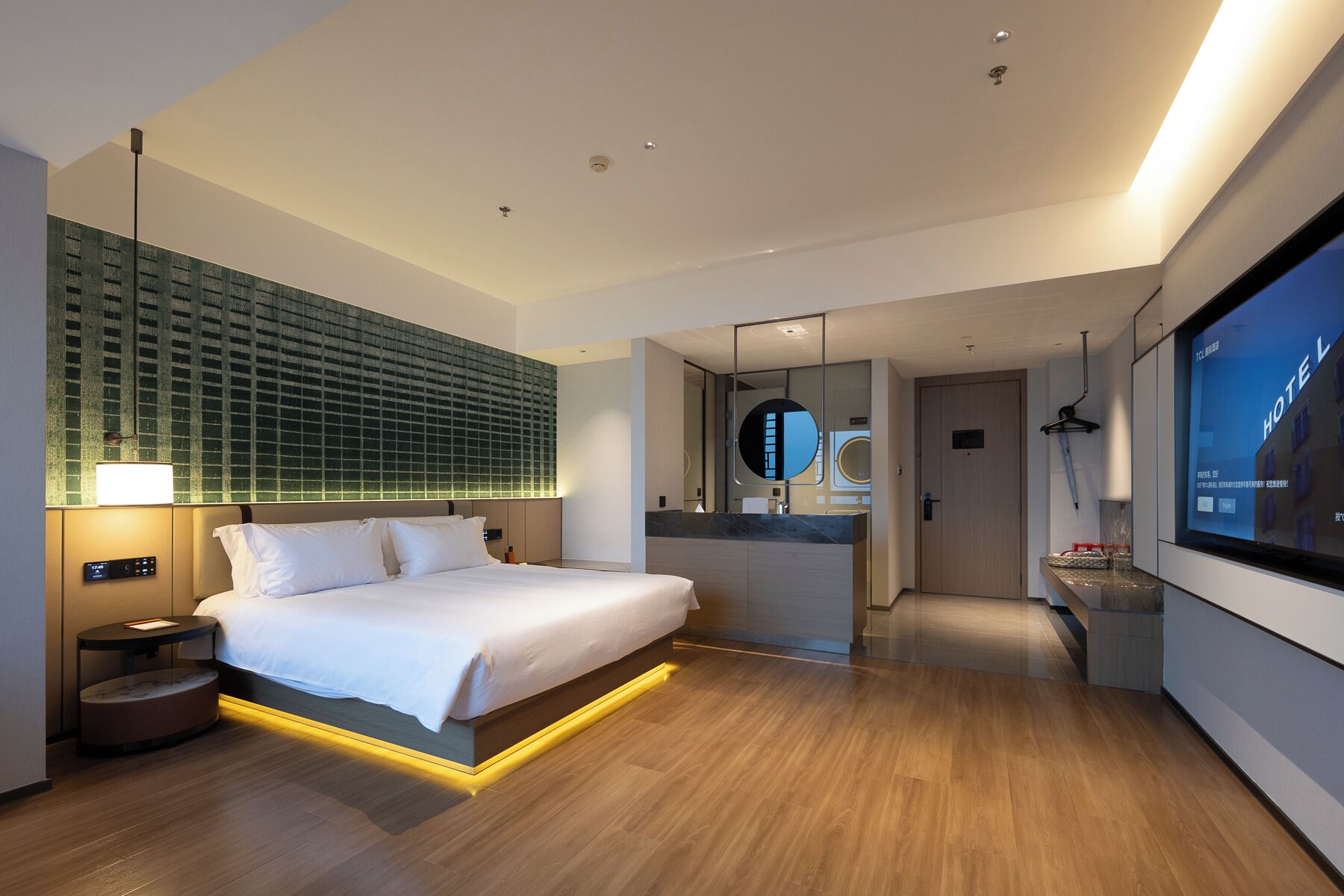 Ein modernes Hotelzimmer mit einem großen Bett, einem Schminktisch mit Spiegel und einem Fernseher, der 'HOTEL' anzeigt.