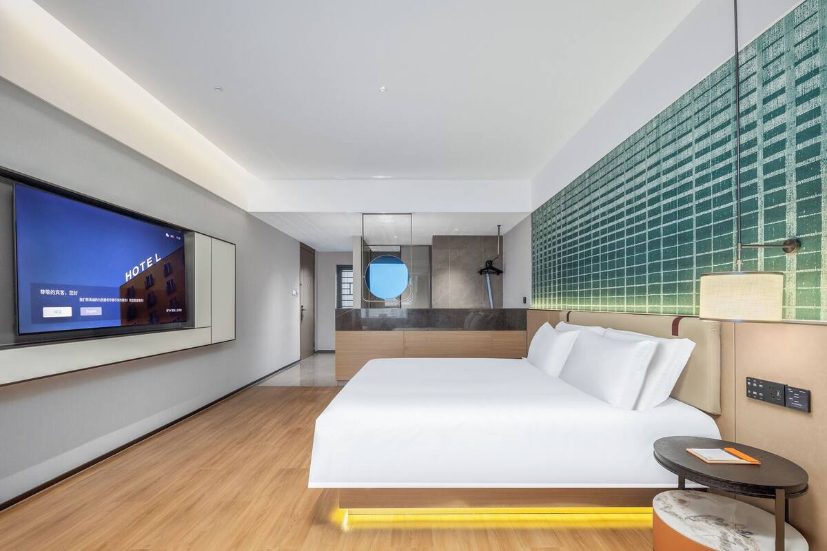 Une chambre d’hôtel moderne avec un grand lit, un téléviseur fixé au mur, une petite table et un mur recouvert de carreaux verts originaux.