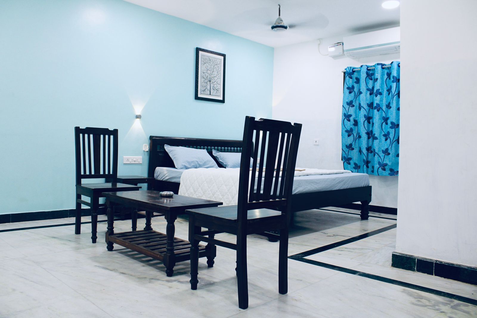 A bedroom with a bed, two chairs, a nightstand, and a wall-mounted air conditioner.