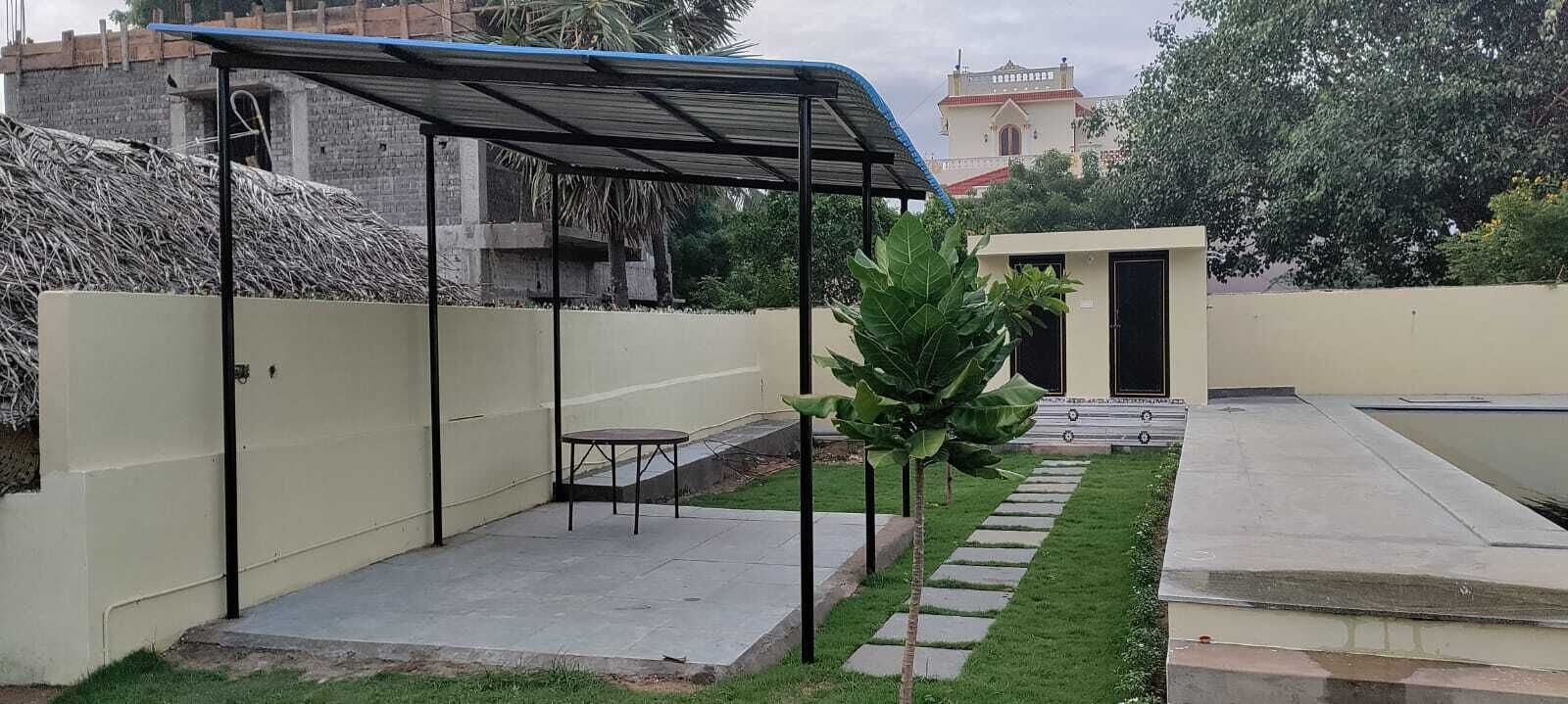 A covered outdoor seating area with a table and chairs, a small garden with a pathway, and a building with a red roof in the background.