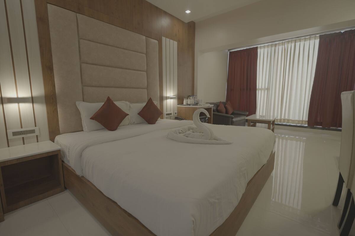 Una habitación de hotel moderna con una cama grande, cabecera de madera, una mesita de noche, un escritorio pequeño y una ventana con cortinas.
