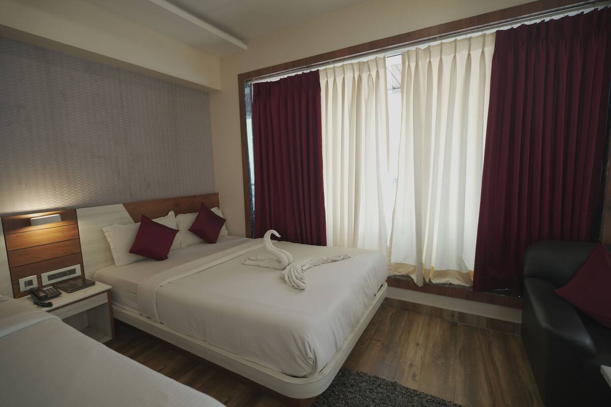 Habitación de hotel con una cama grande, una mesita de noche con lámpara, cabecera de madera y vistas al exterior a través de una ventana con cortinas rojas.