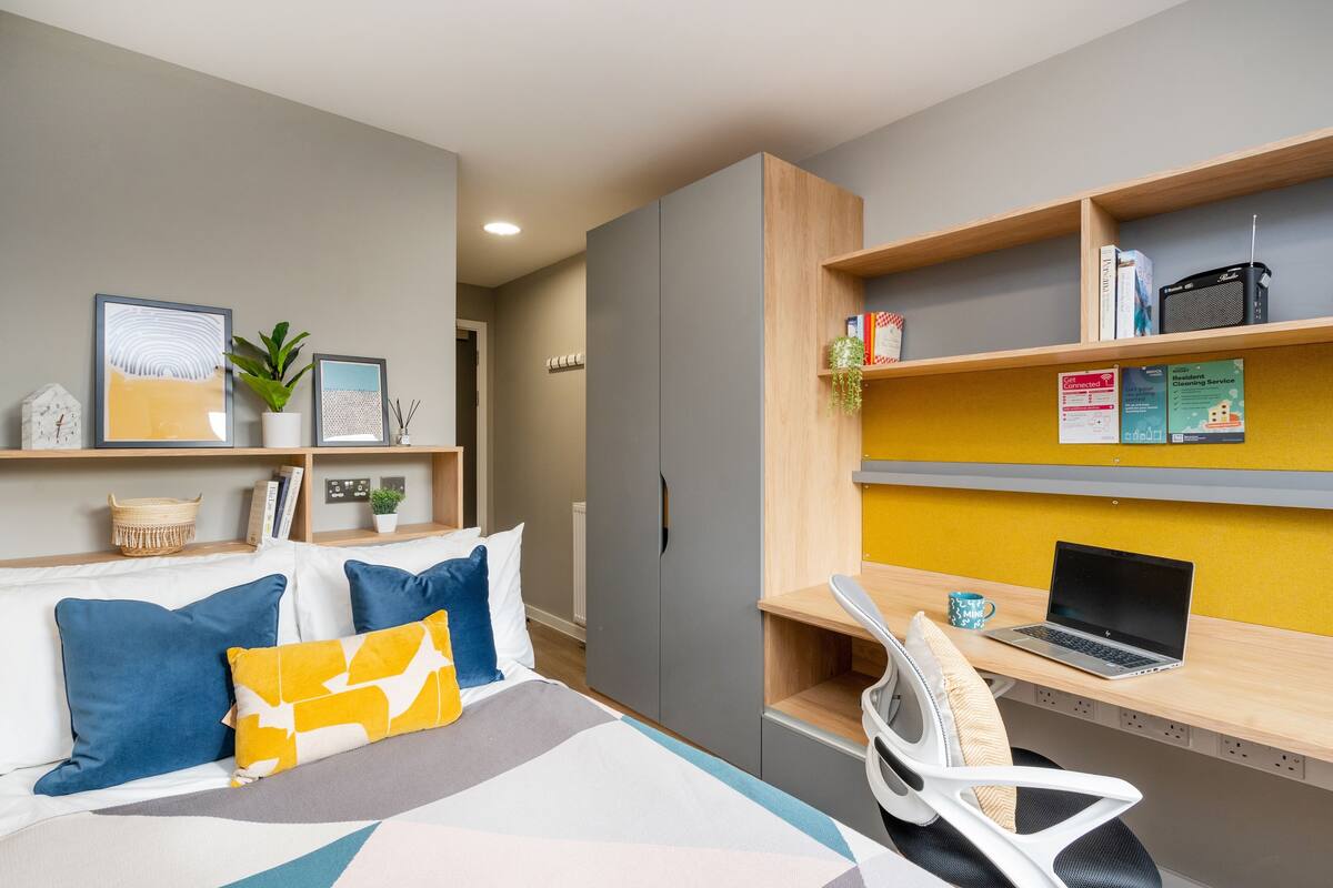 A modern hotel room with a bed, a desk with a laptop, shelves with books and a plant, and a wall-mounted shelf with a framed picture.