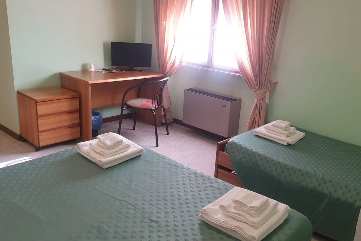 A hotel room with two beds, a desk with a computer, a chair, a radiator, and a window with curtains.