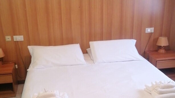 Una cama bien hecha con sábanas y toallas blancas, un cabecero de madera y una mesita de noche con una lámpara.