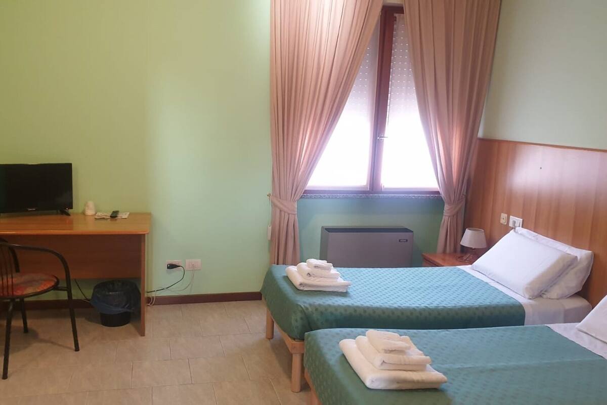 A small hotel room with two single beds, a desk with a chair, a television, and a window with curtains.