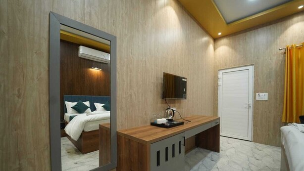 A hotel room with a wooden wall, a bed with green and white bedding, a wooden desk with a TV, and a white door. - Brij Hari Villa (Varanasi)