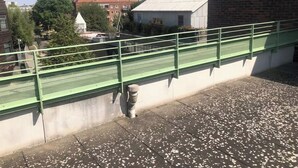 Eine Dachterrasse mit grüner Metallreling, einem Betonboden mit weißen Tupfen und einem Backsteinhaus im Hintergrund.