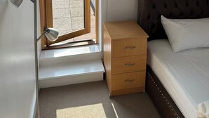 Ein Schlafzimmer mit einem Bett, einem kleinen Nachttisch, einer Schreibtischlampe und einem Fenster mit Blick auf eine Stadtlandschaft.