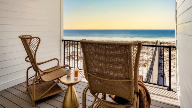 Ein Balkon mit Korbstühlen und einem kleinen Tisch, der Blick auf einen Strand mit freier Sicht auf das Meer und den Sand bietet.