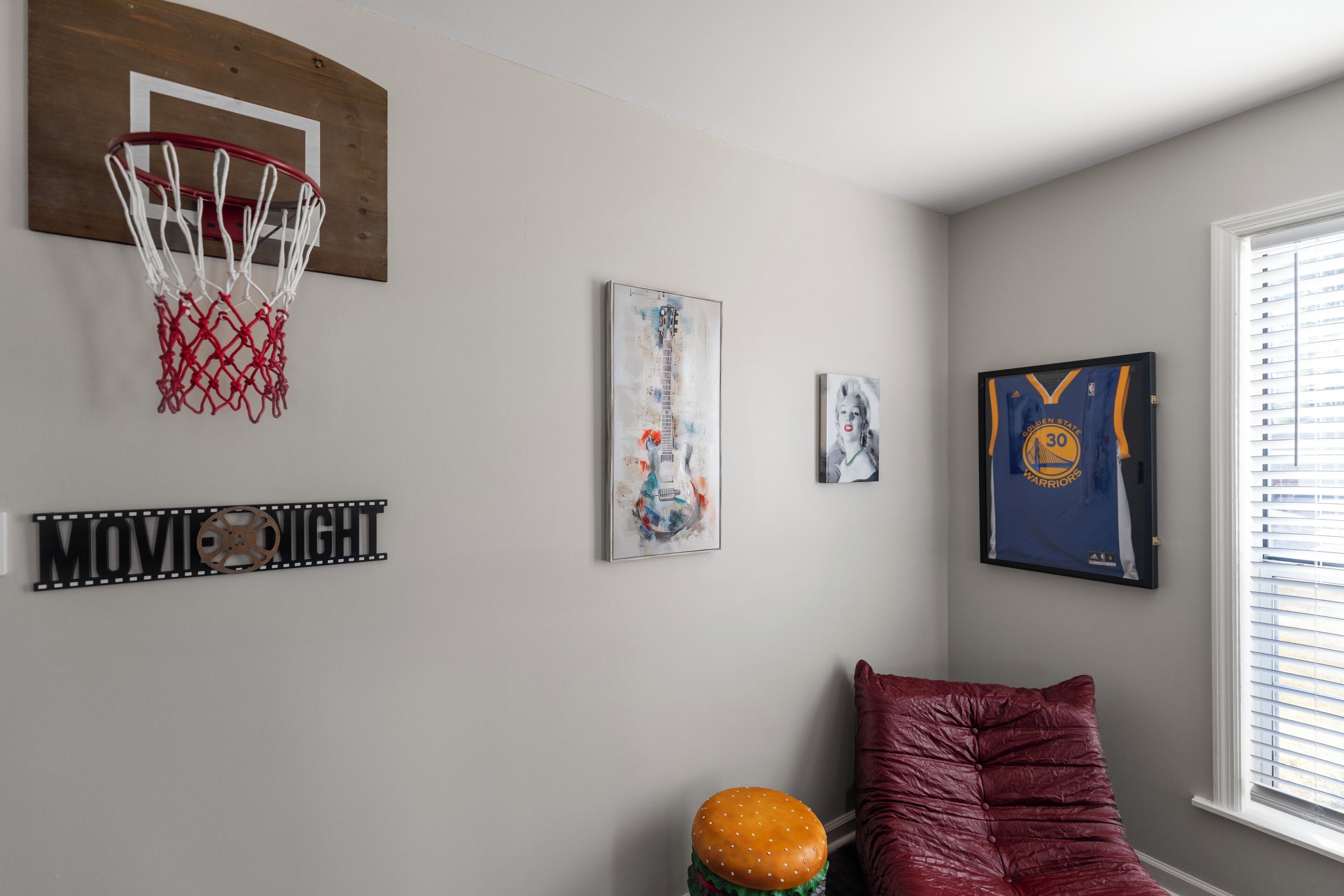 Une pièce avec un panier de basket, une affiche encadrée, un fauteuil rouge moelleux et une fenêtre avec des stores.