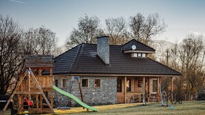 Una casa con fachada de piedra, una estructura de juegos de madera y un tobogán verde.