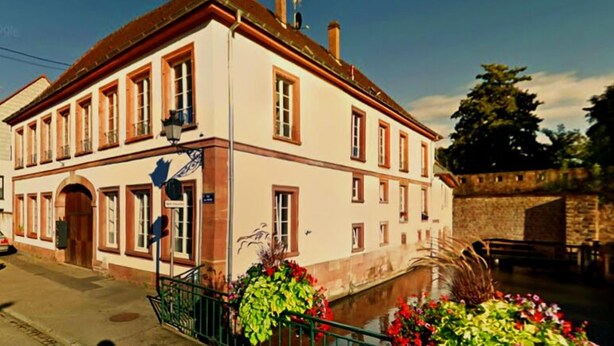 A two-story building with a white facade and red trim, featuring a large arched entrance and multiple windows. - Les gîtes de Wissembourg- Color Lodge (Wissembourg)