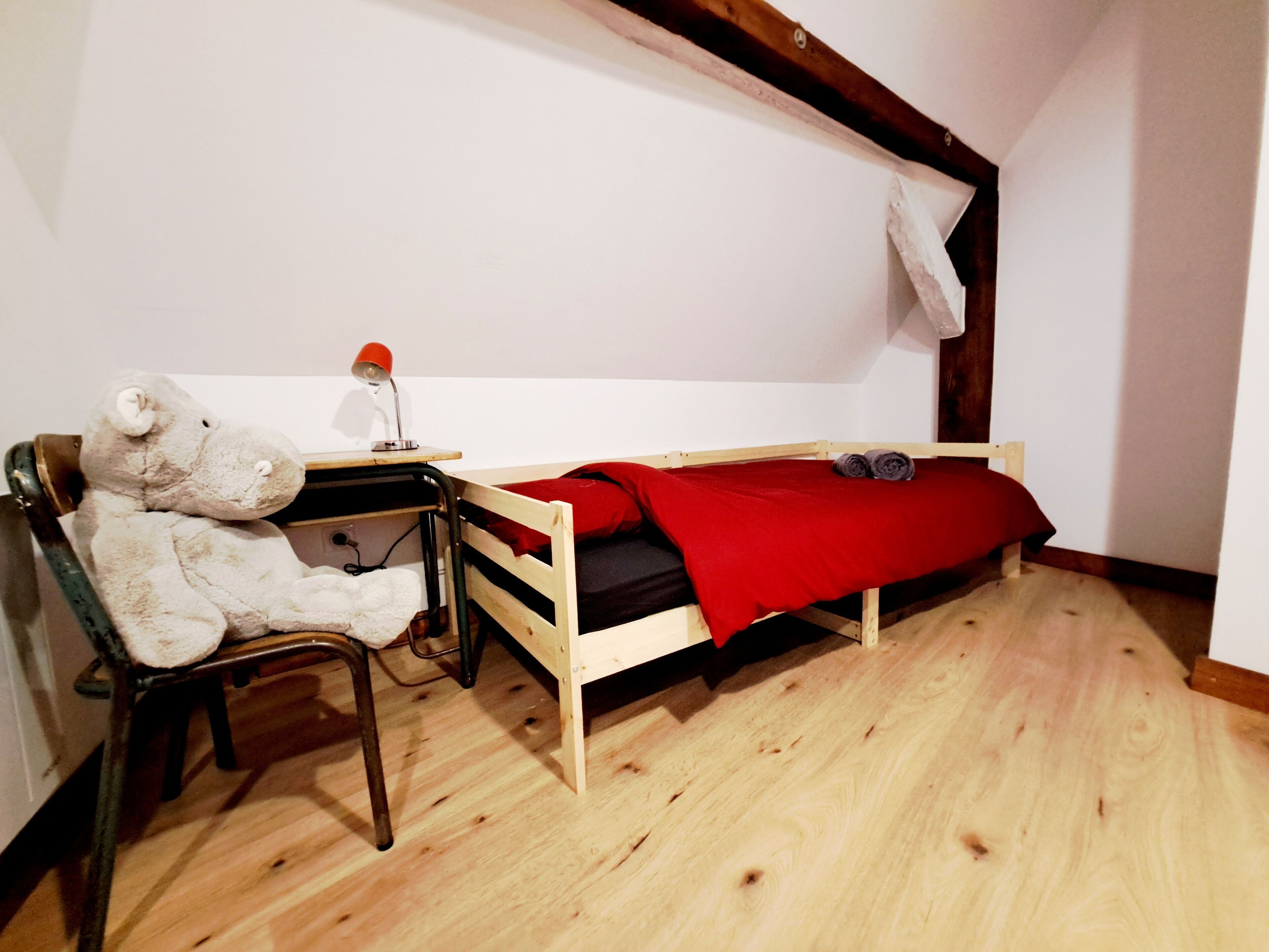 A small, single-room accommodation with a wooden floor, a bed with a red blanket, a desk with a chair, and a teddy bear.