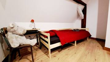 A small, single-room accommodation with a wooden floor, a bed with a red blanket, a desk with a chair, and a teddy bear.