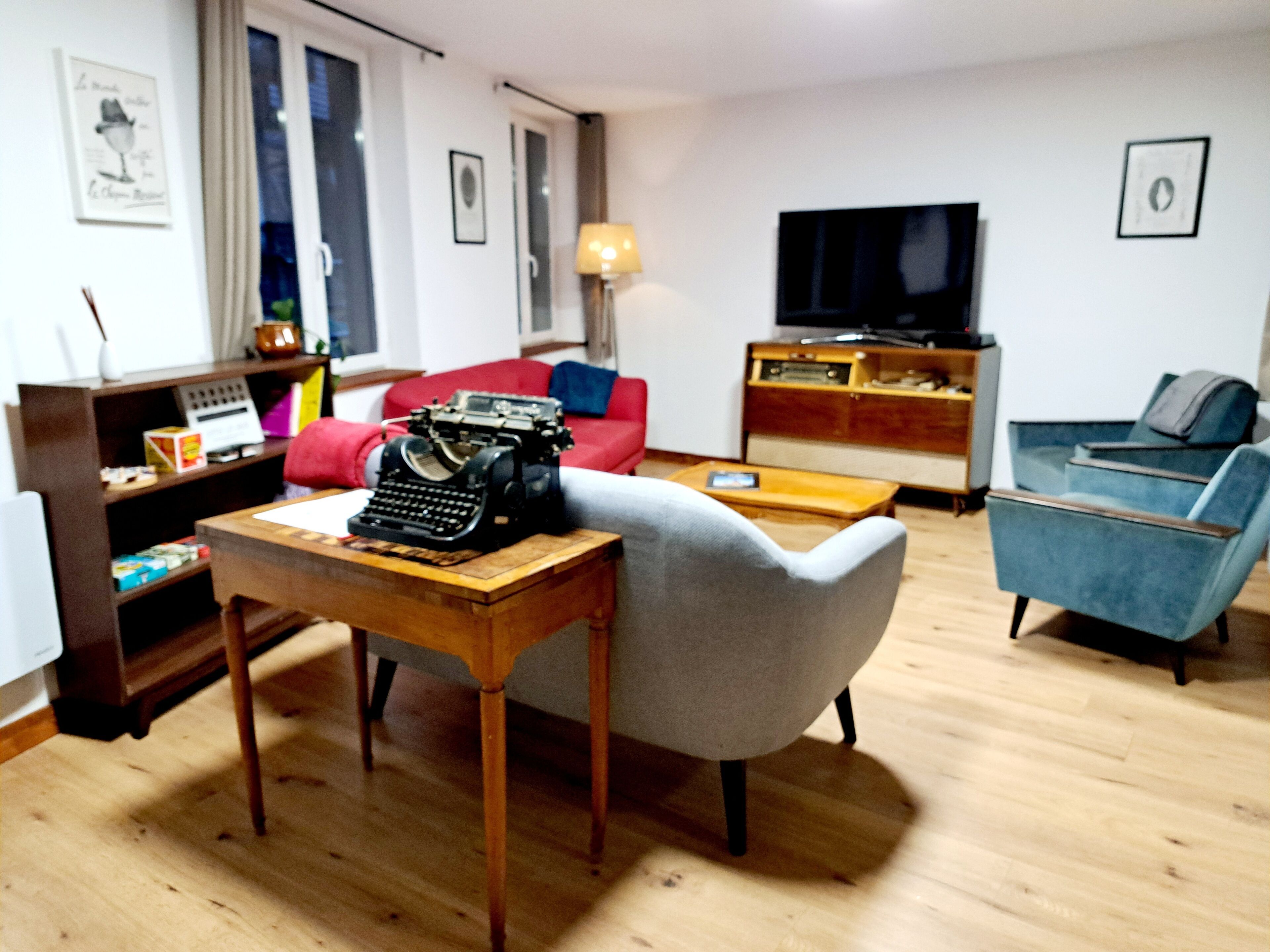 Ein Wohnzimmer mit einem Holztisch, einem roten Sofa, einem blauen Sessel, einem Fernseher und einem Bücherregal.