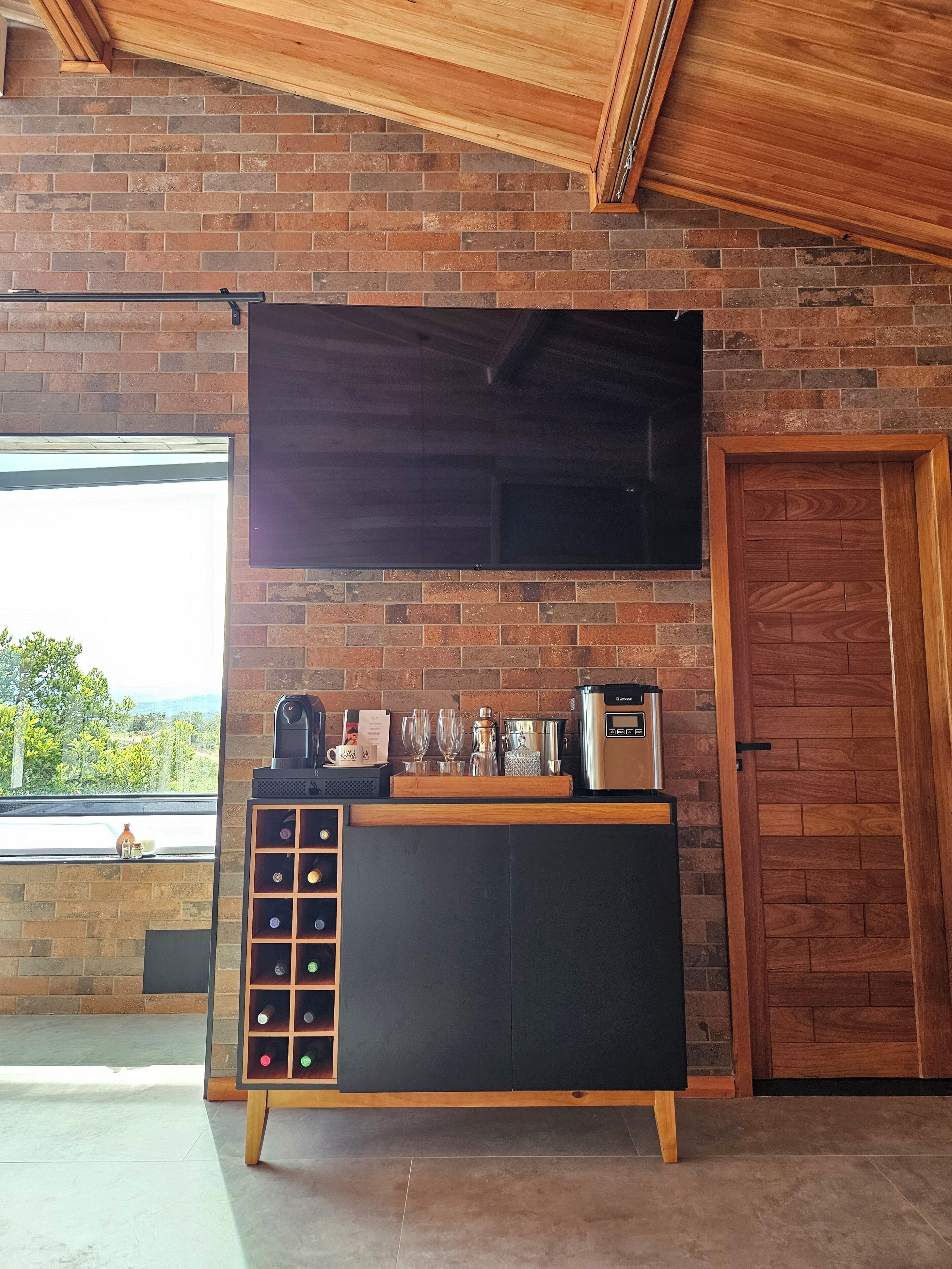 A modern kitchen with a brick wall, a large flat-screen TV, a wine rack, and a coffee machine.