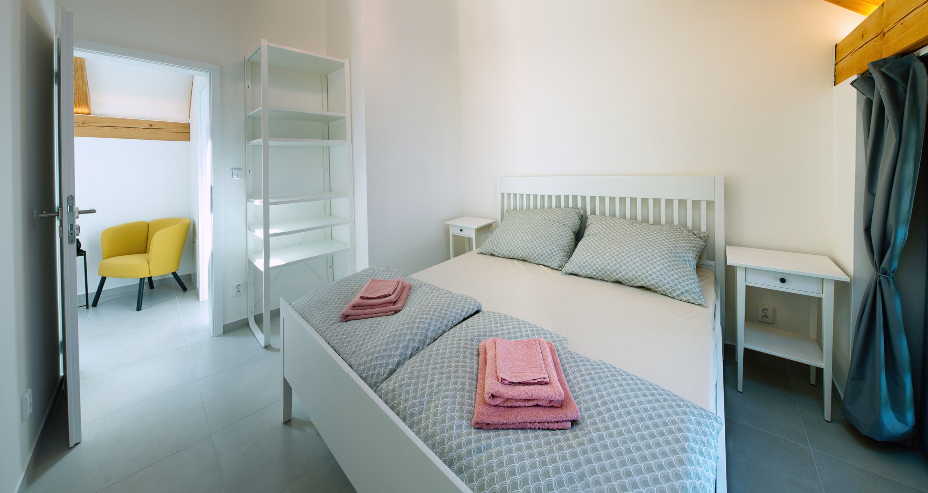 A bedroom with a bed, two bedside tables, a chair, a wardrobe, and a door leading to another room.
