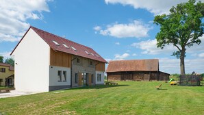Ein modernes Haus mit geneigtem Dach, ein großer Baum und ein Bauernhof im Hintergrund.