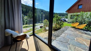 Ein Zimmer mit großem Fenster mit Blick auf eine Steinterrasse und einen bewaldeten Bereich.
