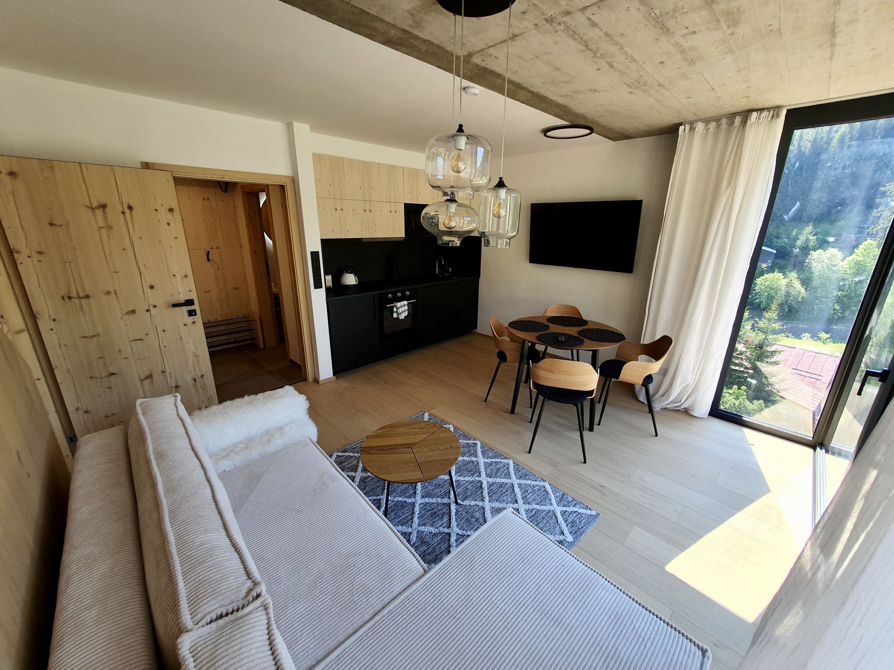 Uma sala moderna com um sofá, uma mesa de jantar com cadeiras, área de cozinha e uma janela grande com vista para o verde.