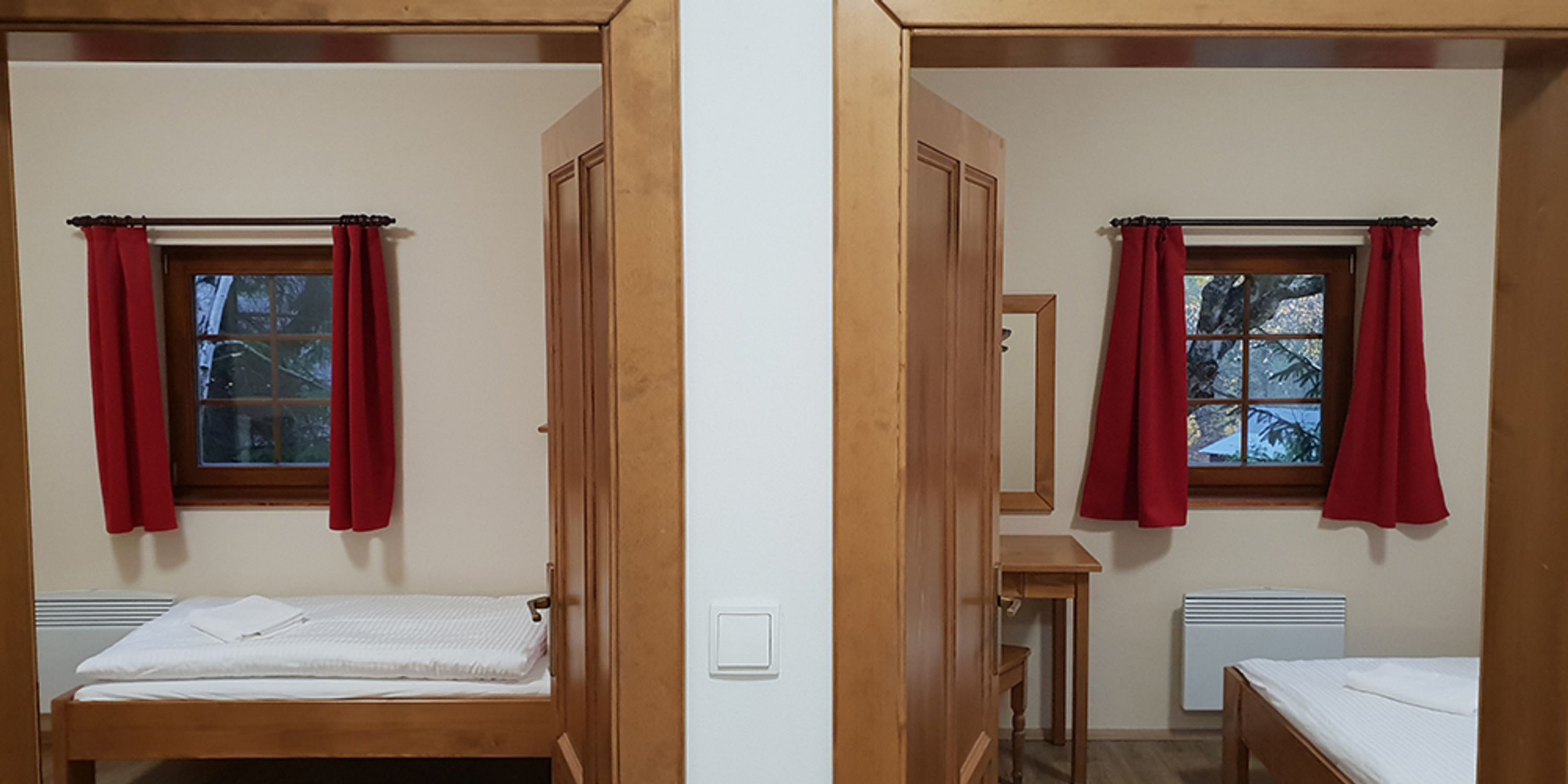 Una habitación con cama, una mesita, un ventanal con cortinas rojas y un radiador.