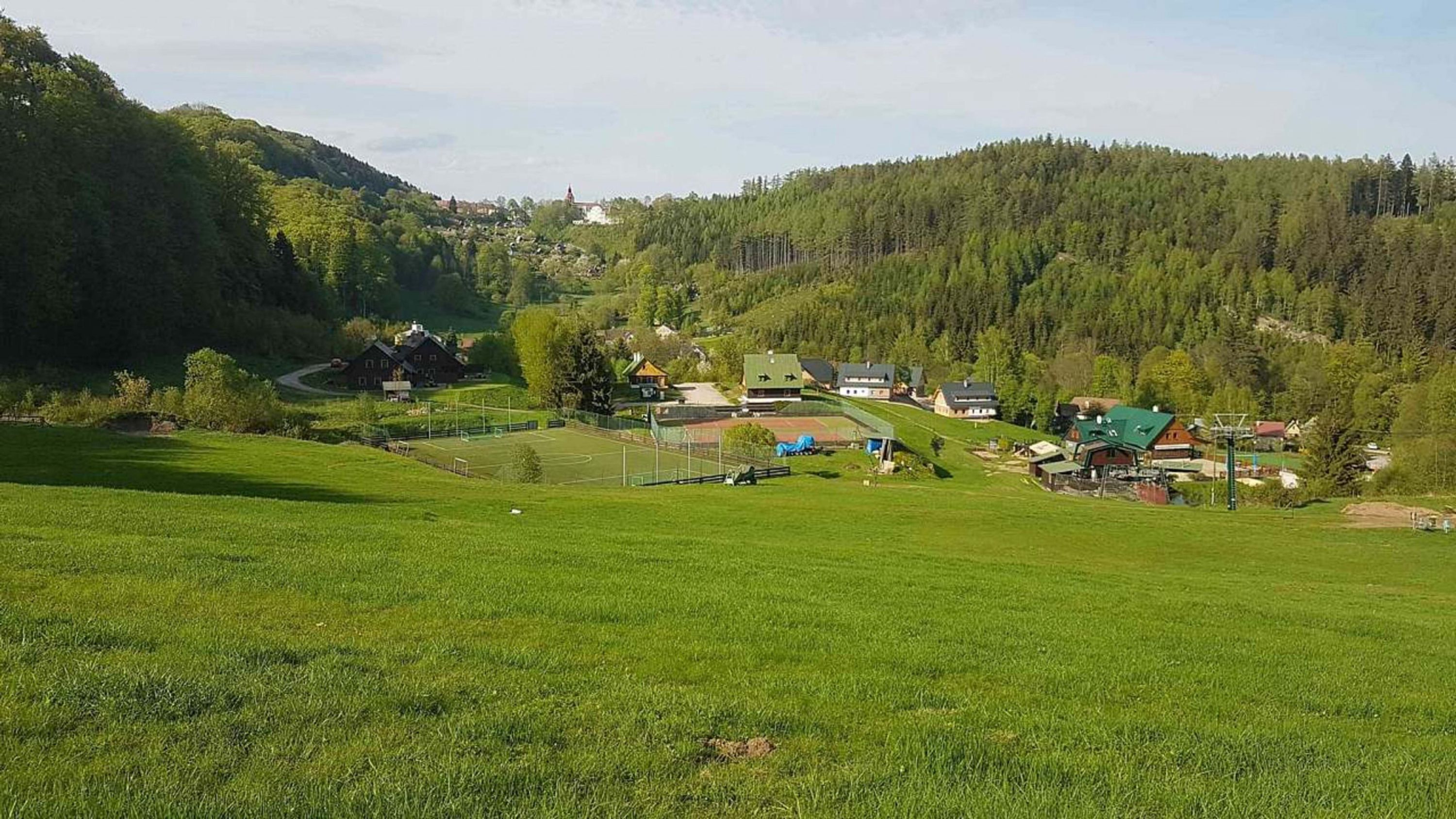Ett lantligt landskap med en grön åker, en liten byggnad och en skogbeklädd kulle i bakgrunden.