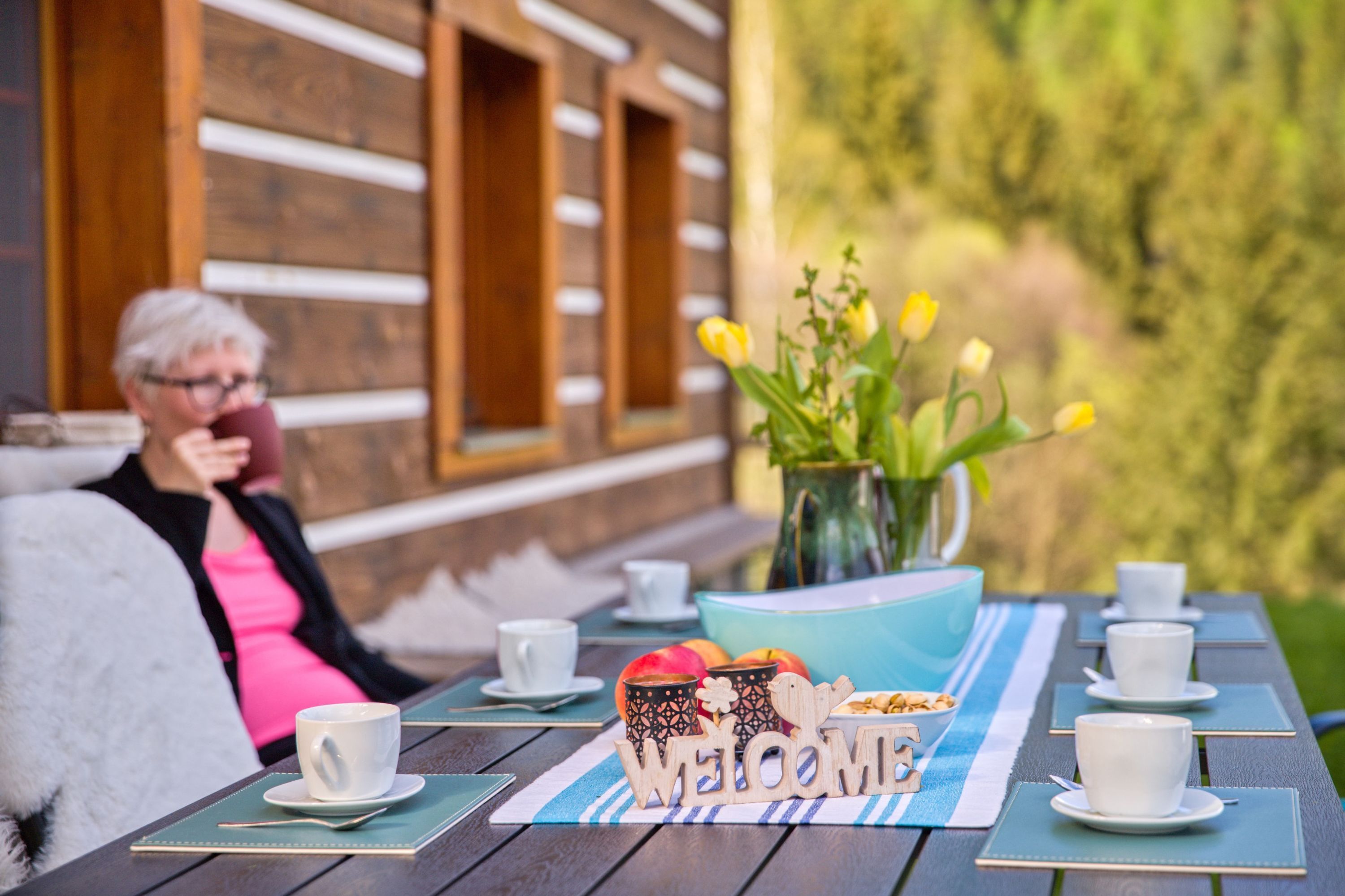 Une terrasse en bois avec une table dressée pour un repas, un panneau d’accueil et un vase rempli de fleurs.