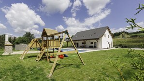 A wooden swing set in a grassy yard with a house in the background. - Prosence Kpa250 in Prosecn (Prosečné)