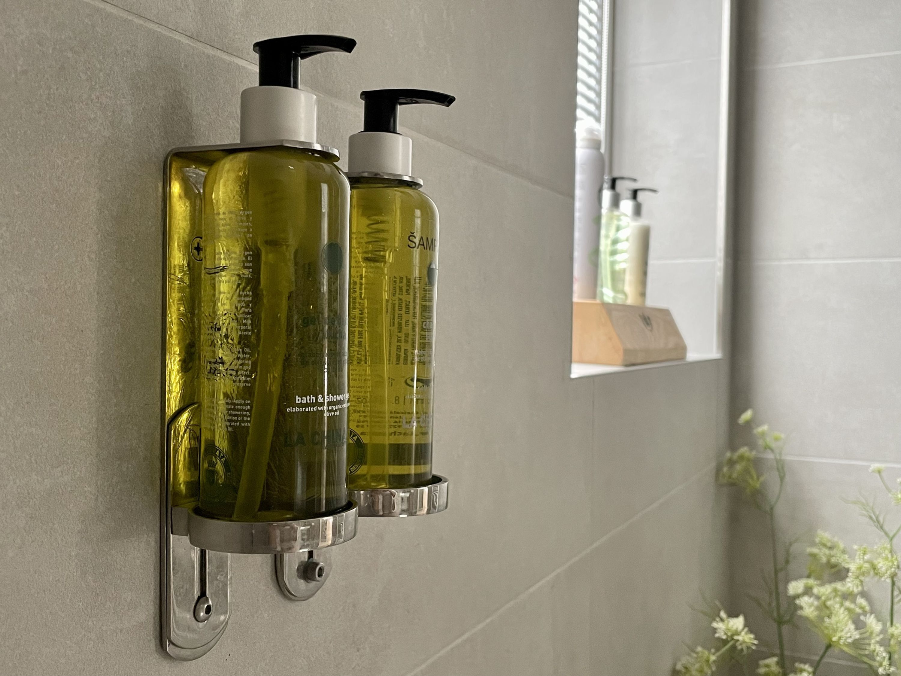 Two bathroom dispensers mounted on a wall, with a window and a plant visible in the background.