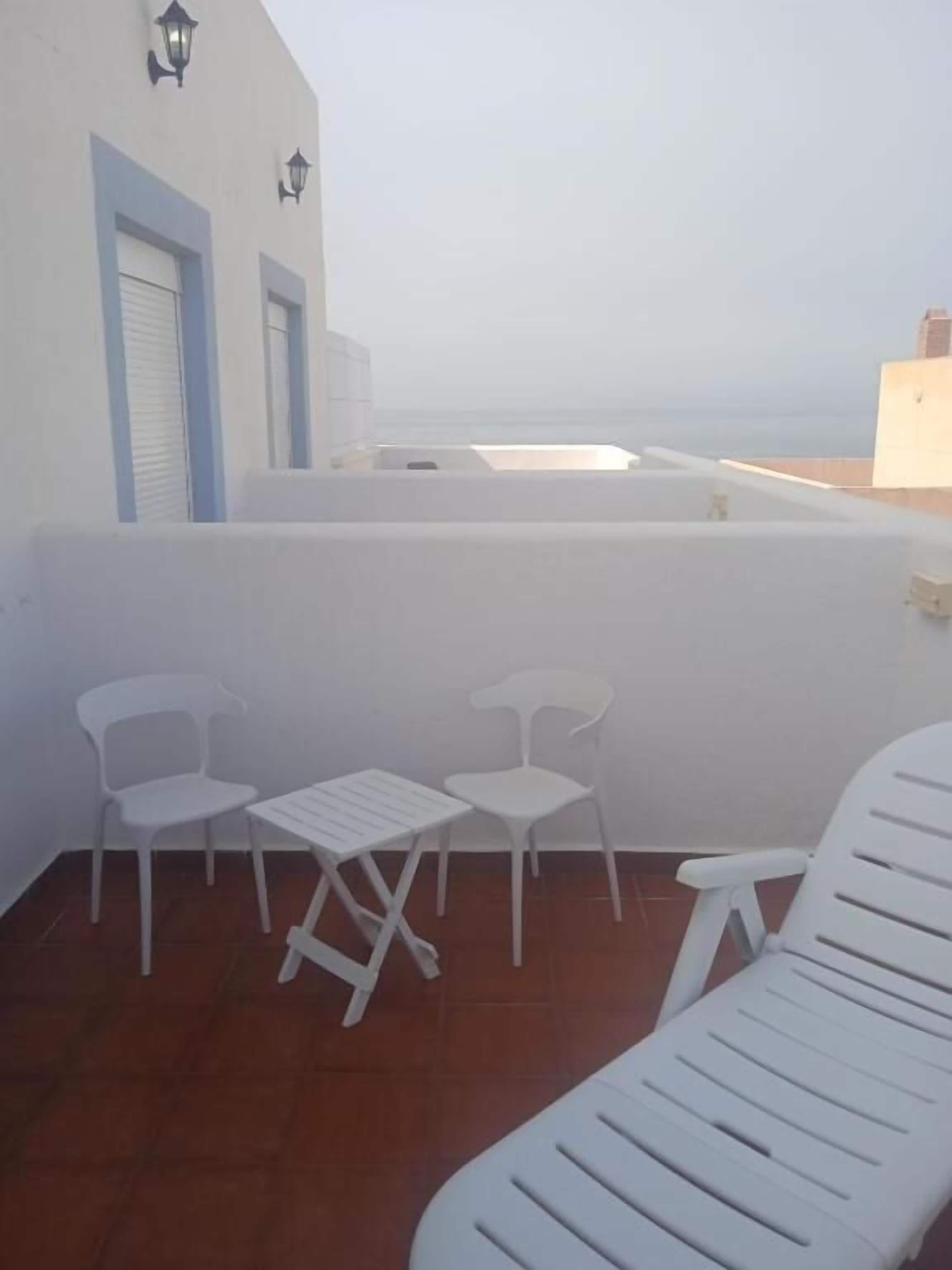 A terrace with white plastic chairs and a table, overlooking a beach.