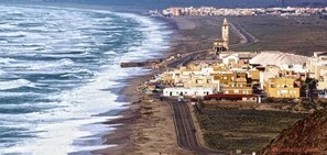 A coastal town with buildings along the beach, a prominent tower, and a road leading to the sea. - HOTEL LAS SALINAS  CABO DE GATA (Almeria)