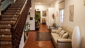 Eine Treppe mit aufwendiger Geländerführung führt zu einer oberen Etage, einem Wohnbereich mit Sofa und einer großen Vase, sowie einem an der Wand befestigten Feuerlöscher.