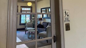 Une porte ornée d’une plaque décorative en forme de blason, d’un panneau indiquant les consignes de sécurité incendie, et une vue sur un salon avec un ventilateur de plafond, un canapé et un fauteuil à motifs.