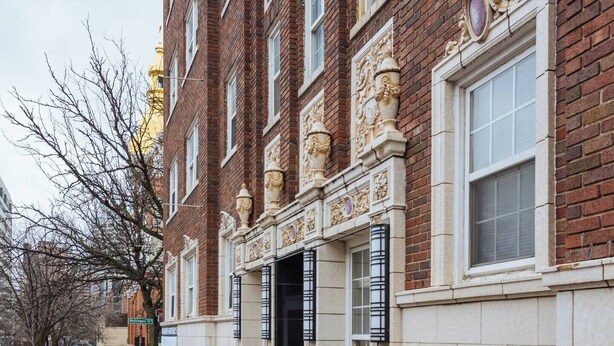 Un bâtiment en briques dont la façade est recouverte de carreaux blancs, avec une porte d’entrée noire et un trottoir devant.