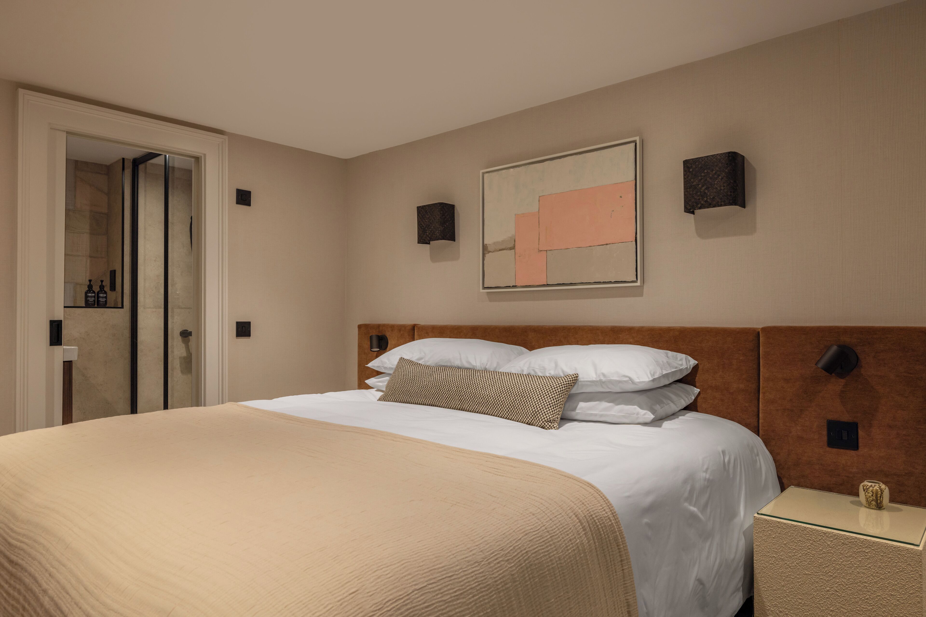 Quarto de hotel com uma cama grande, box de vidro para o chuveiro, uma obra de arte na parede e uma pequena mesa de cabeceira com um vaso.