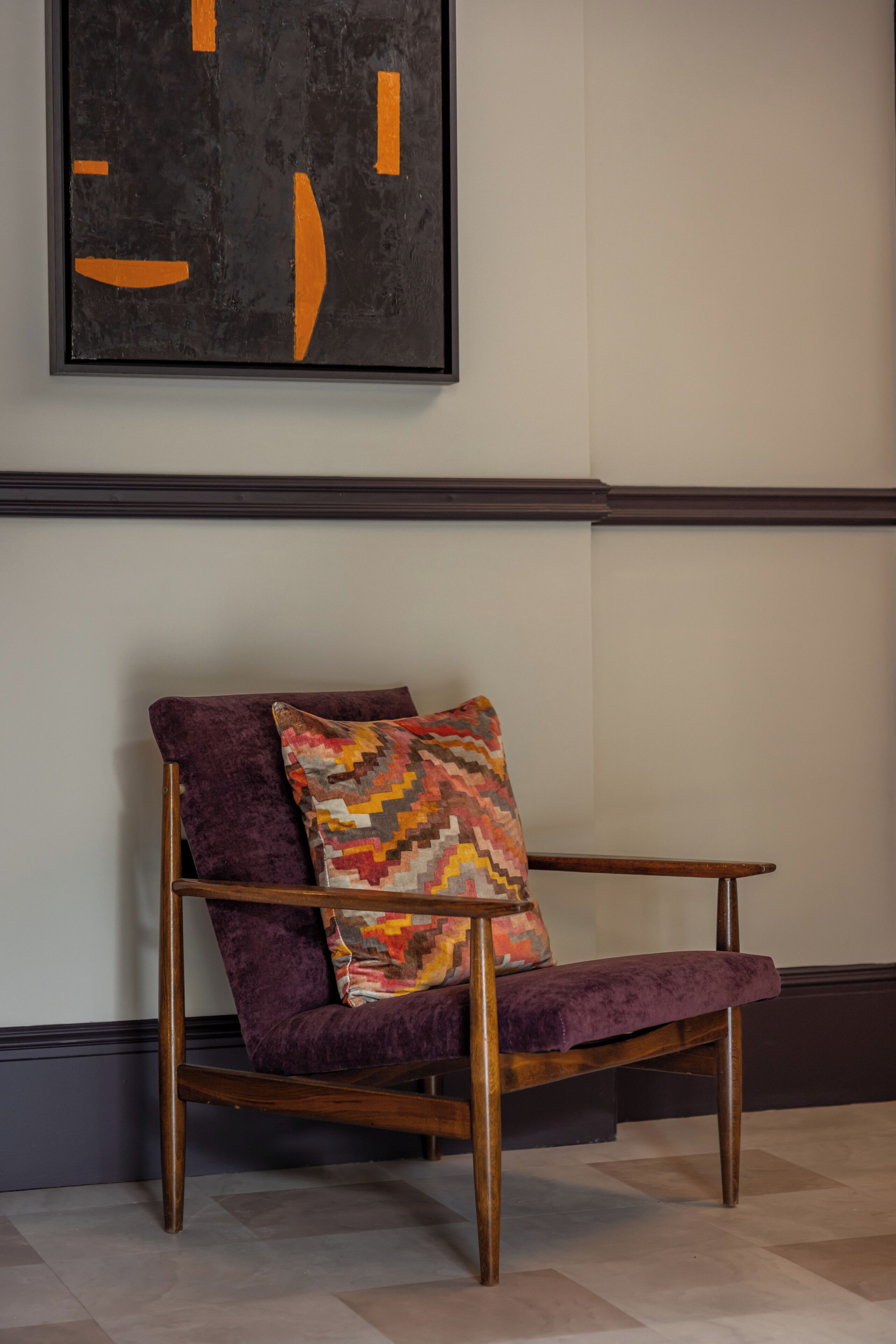 A chair with a purple cushion and a colorful geometric pillow, positioned in front of a wall with a black and orange abstract painting.