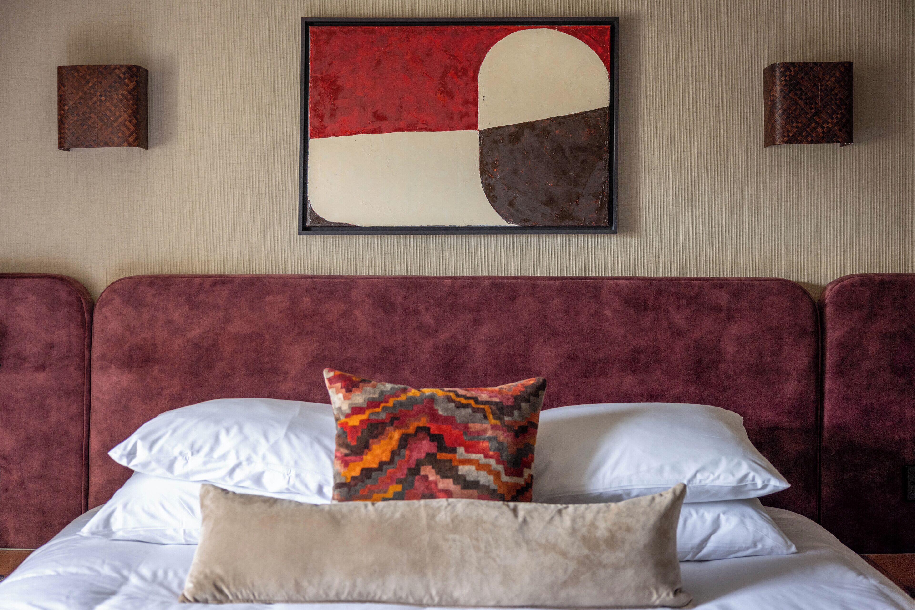 Ein Bett mit weißen und beigen Kissen, einem farbenfrohen geometrischen Kissen sowie einem großen abstrakten Gemälde an der Wand.