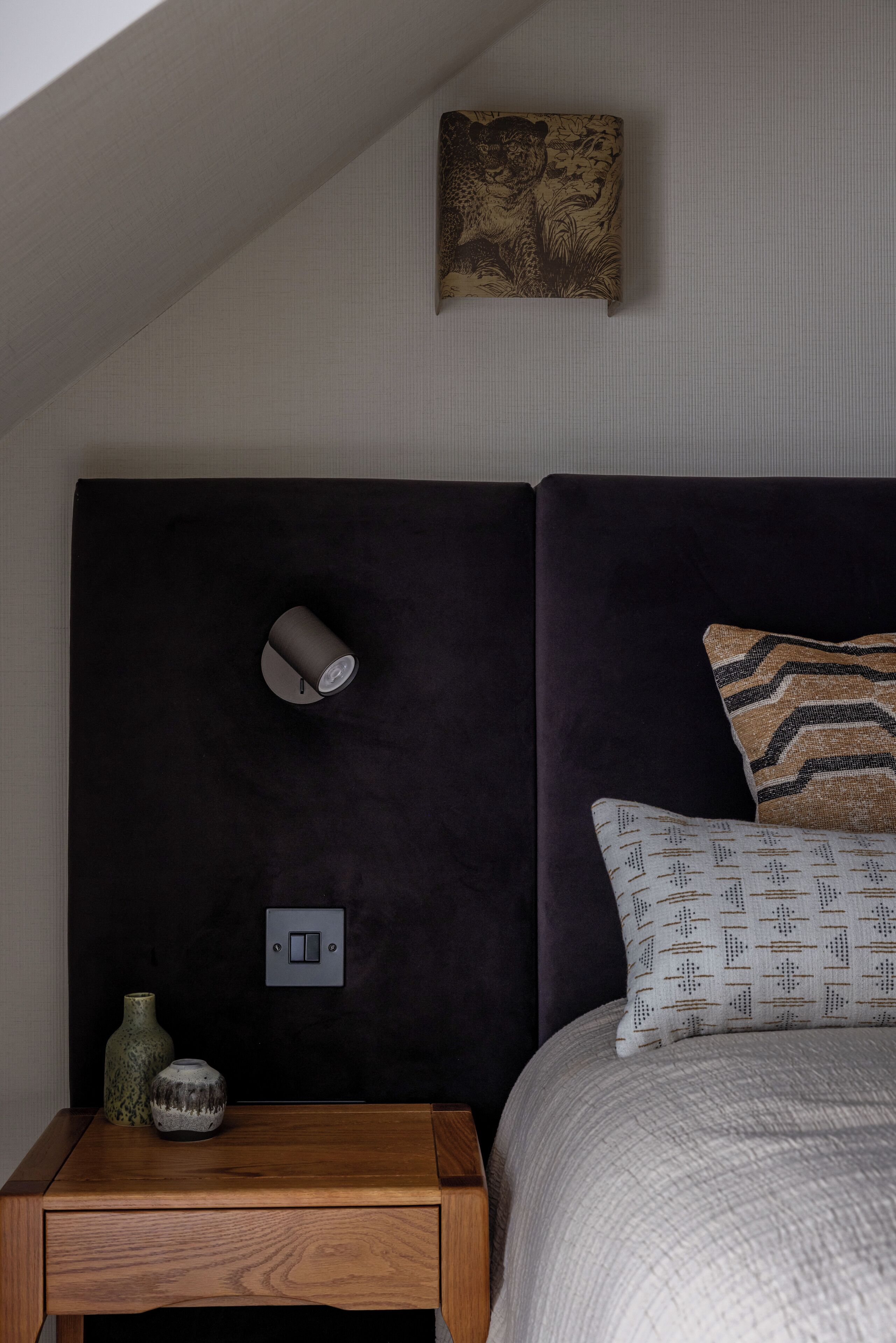 A bedroom with a dark headboard, a bedside table with a vase, and a wall-mounted light fixture.